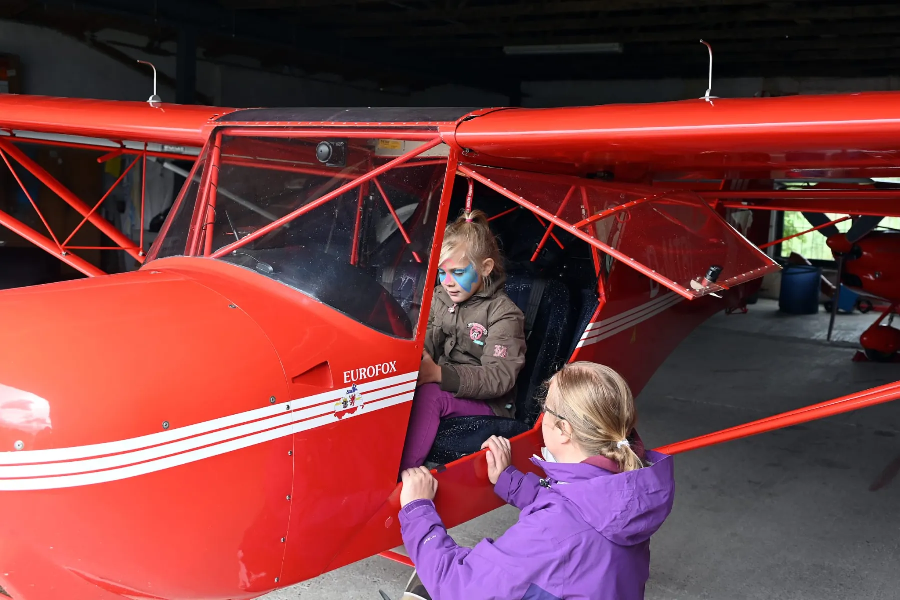Zwei Mädchen klettern in ein rotes Kleinflugzeug im Hangar.