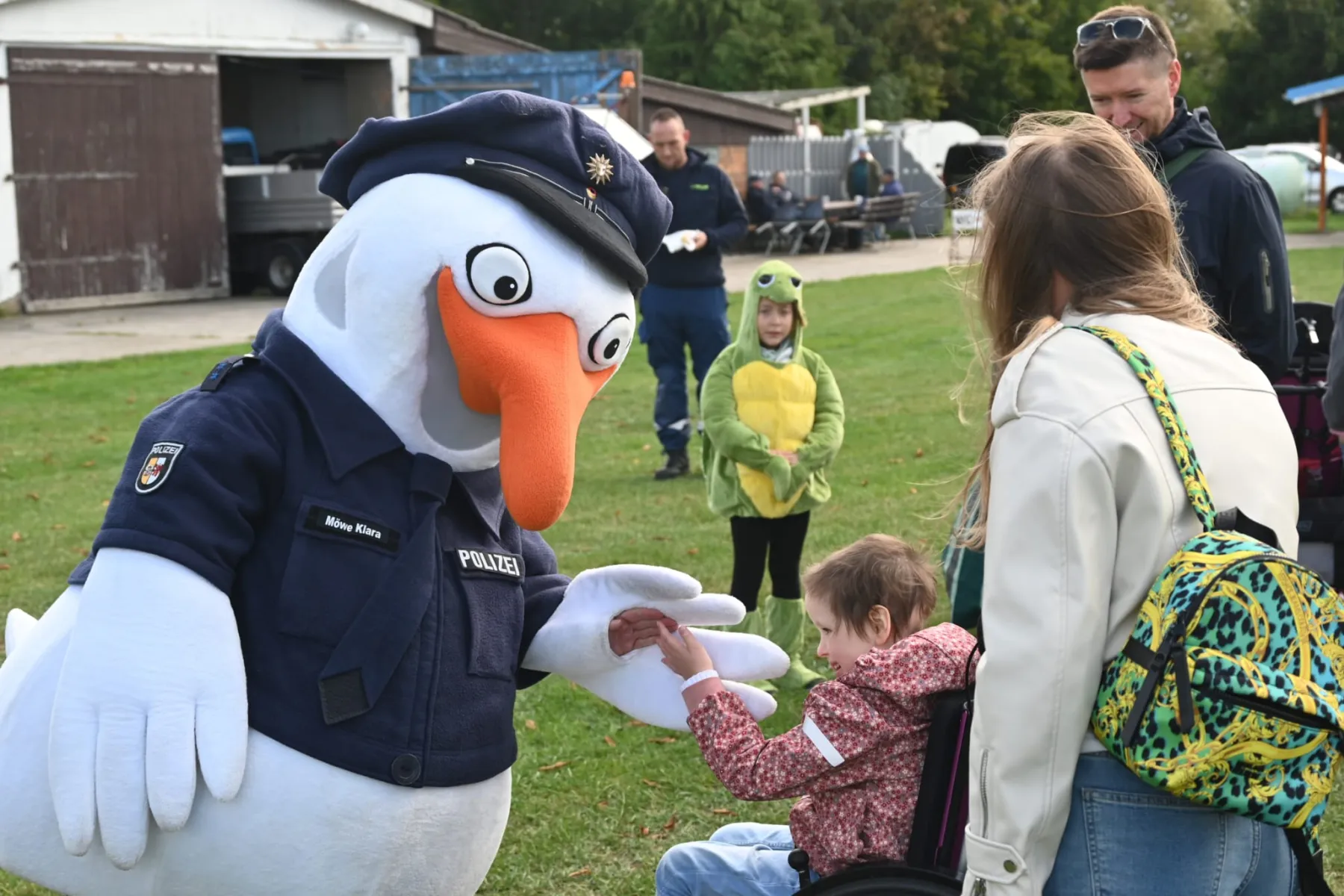 Ein Storch in Polizeiuniform begrüßt ein Kind im Rollstuhl.
